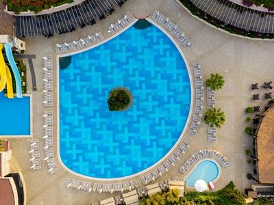 Großer, herzförmiger Pool mit blauem Wasser, umgeben von Sonnenliegen und grünen Pflanzen.