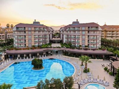 Große Hotelanlage mit zwei Gebäuden, großem blauen Pool und umliegenden Palmen bei Sonnenuntergang.