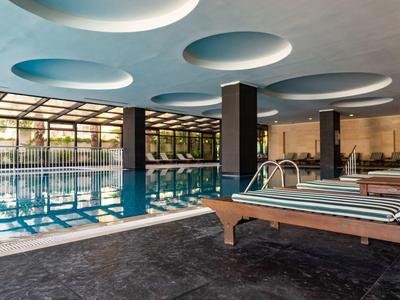 Helles Hallenbad mit Glasfront, blauem Wasser, Saunaliegen und rundem Deckendekor.