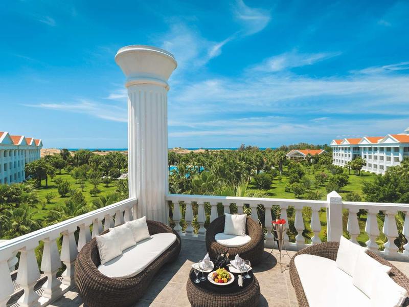 Balcony with seating area overlooking green landscape and buildings under blue sky.
