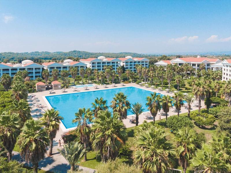 Large hotel resort with a big rectangular pool and many palm trees in sunny weather.