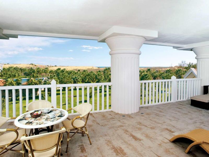 Spacious balcony with white railing, table with chairs, and lounge furniture overlooking green area.
