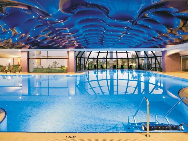 Indoor pool with blue illuminated water and artistically designed ceiling in a hotel.