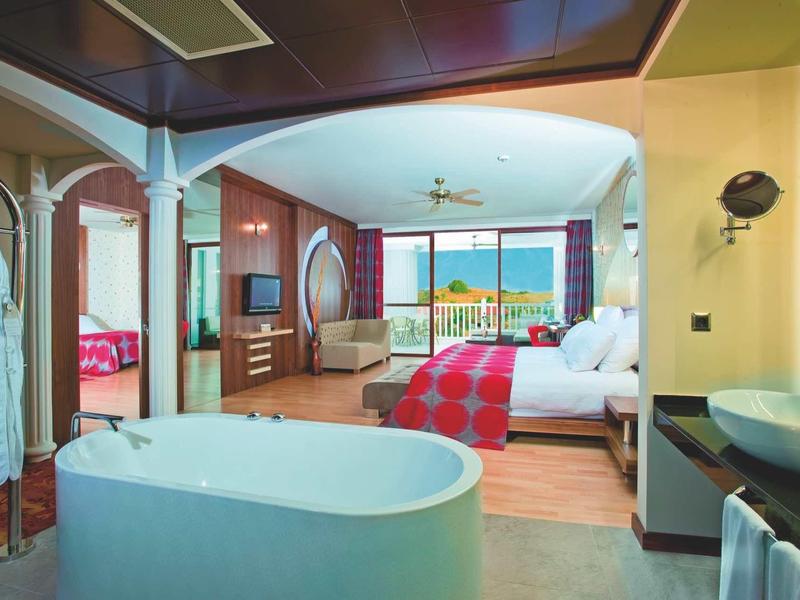 Open hotel room with bathtub, large bed, and sea view from the balcony.