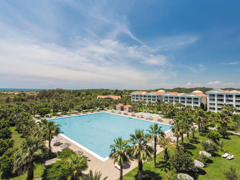 Large hotel pool with palm trees and multi-story building under blue sky.