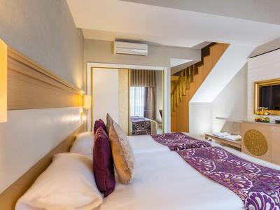 Hell eingerichtetes Hotelzimmer mit großem Bett, lila Bettläufer, Holztreppe und Wand-TV.