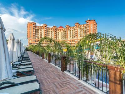 Hotel mit mehrstöckigem Gebäude, Pool, Palmen und Liegestühlen an Sonnenschirmen auf Terrasse.