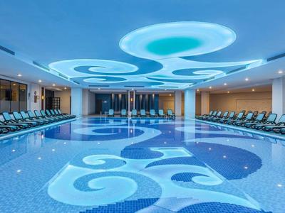 Großes Hallenbad mit türkisblauem Wasser, modernen Liegestühlen und heller Deckenbeleuchtung.
