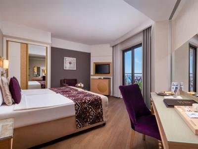 Modern eingerichtetes Hotelzimmer mit Doppelbett, Schreibtisch, lila Akzenten und großem Fenster.