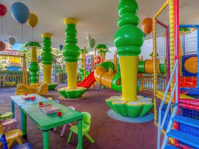 Area giochi interna colorata con strutture per arrampicarsi, scivoli e piccoli tavoli con sedie.