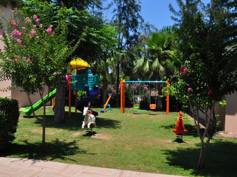 Parco giochi per bambini con scivolo, altalene e struttura da arrampicata in un giardino con alberi.