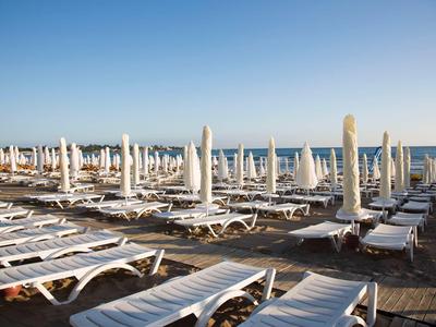Viele weiße Liegestühle und geschlossene Sonnenschirme stehen dicht am Strand vor blauem Himmel.