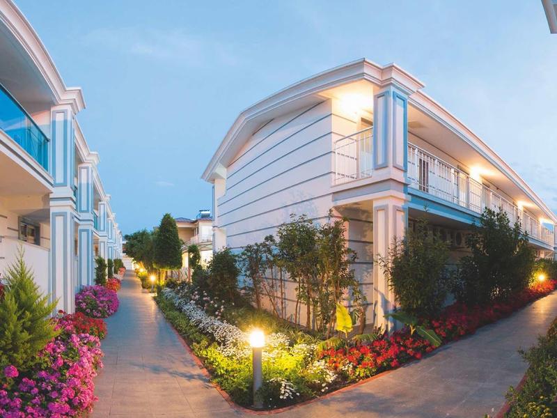 Zwei weiße Häuser mit Blumenbeeten und beleuchtetem Weg bei Dämmerung unter blauem Himmel.