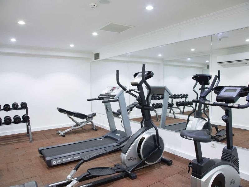 Heller Fitnessraum mit Laufbändern, Fahrradergometern, Gewichten und großen Spiegeln an der Wand.