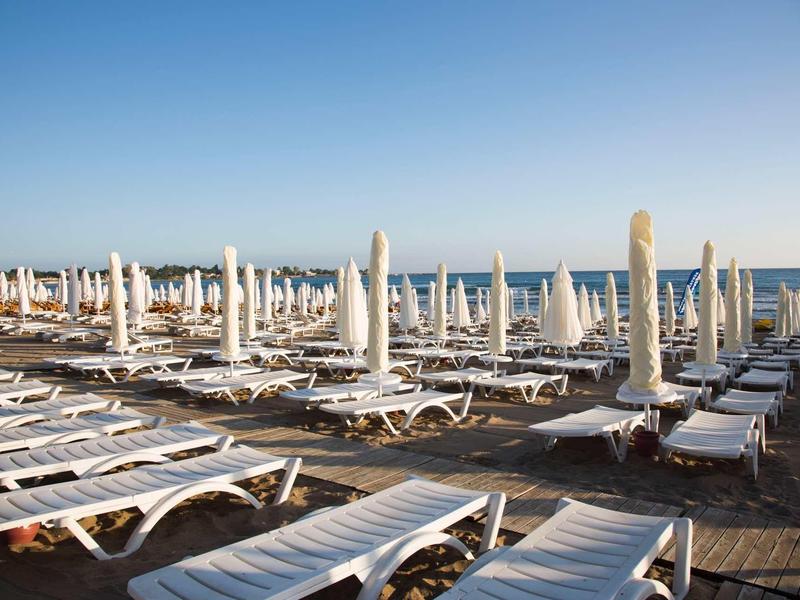 Viele weiße Liegestühle und geschlossene Sonnenschirme stehen dicht am Strand vor blauem Himmel.