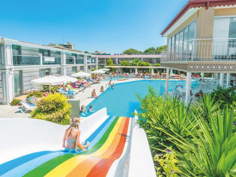 Outdoor-Pool mit bunter Rutsche, umgeben von Liegestühlen und modernem Hotelgebäude.
