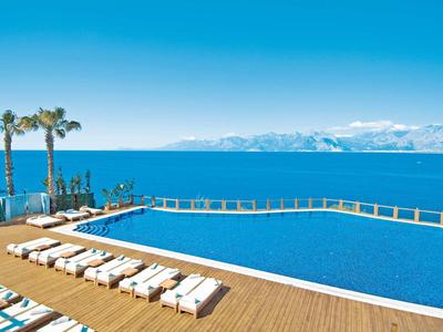 Großer Infinity-Pool mit Holzdeck, Liegestühlen und Palmen vor blauem Meer und Bergen im Hintergrund.