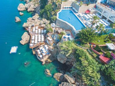 Küste mit Felsen, Pool und mehreren Sonnenschirmen bei klarem blauem Wasser und grünem Bewuchs.