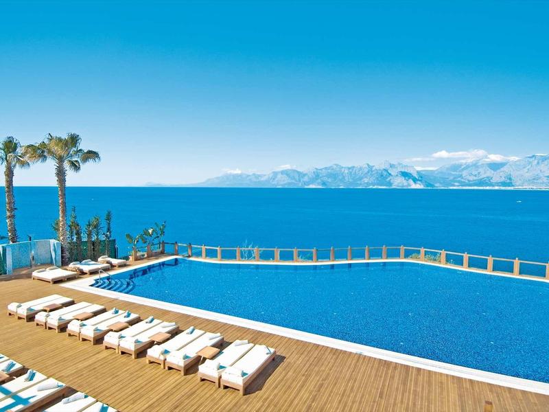 Groot zwembad met ligstoelen op houten dek met uitzicht op blauwe zee en bergen.