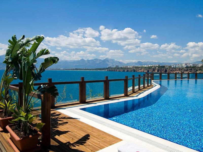 Infinity zwembad met houten vlonder en planten naast zee en bergen onder een heldere lucht