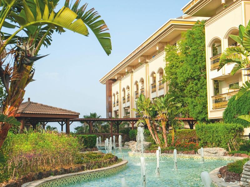 Hotel con giardino lussureggiante, fontane e piante tropicali sotto un cielo azzurro.