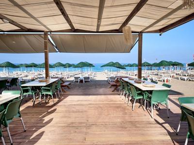 Offene Terrasse mit grünen Tischen und Stühlen mit Blick auf einen Strand mit Sonnenschirmen.