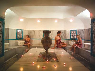 Innenraum eines traditionellen türkischen Bades mit Mosaikfliesen, zentralem Brunnen und drei sitzenden Personen.