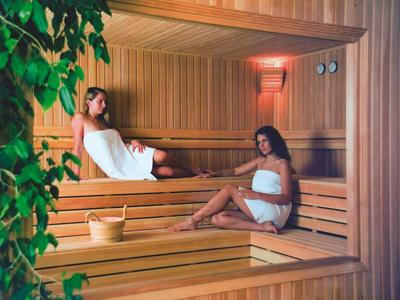 Zwei Frauen in Handtüchern sitzen entspannt in einer hellen Holzsauna mit Eimer und Schöpflöffel.