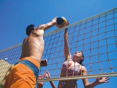 Zwei Personen spielen Beachvolleyball, ein Mann blockt den Ball am Netz bei klarem Himmel.