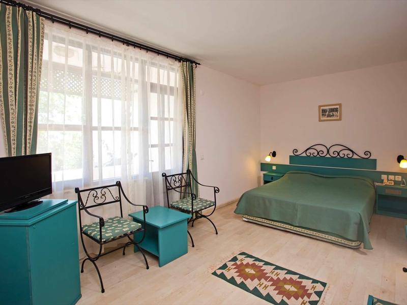 Chambre d'hôtel avec grand lit vert, deux fauteuils en métal et petite table, rideaux aux fenêtres.