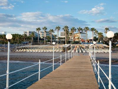 Holzsteg mit weißen Laternen führt zu Palmen und Strandrestaurant unter blauem Himmel.