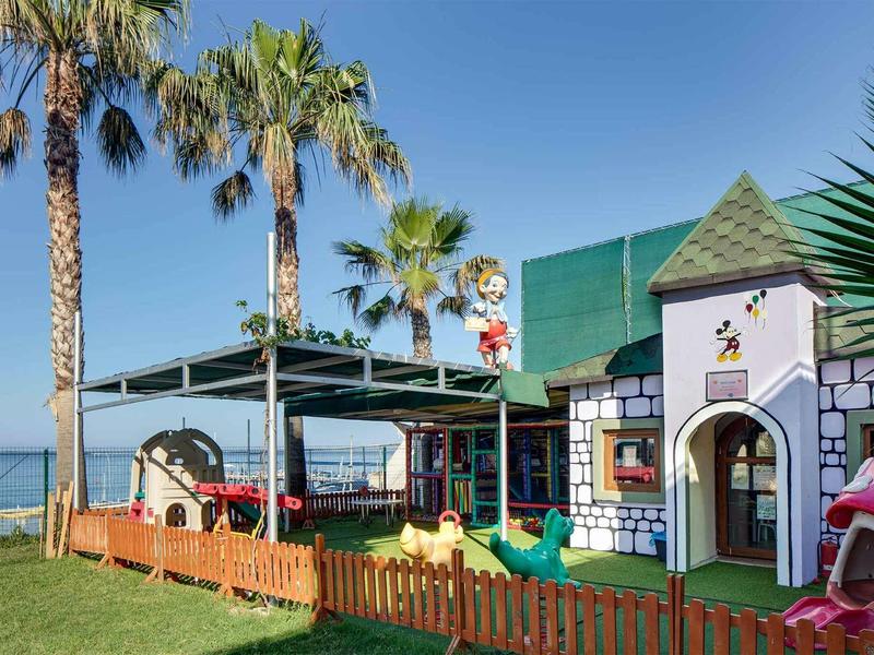 Bunter Kinderspielplatz mit Rutsche, Kletterhaus und Palmen vor klarem blauem Himmel.