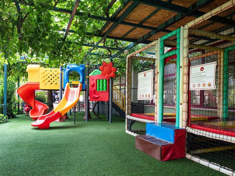 Bunter Kinderspielplatz mit Rutsche, Klettergerüst und gepolstertem Boden unter Bäumen.