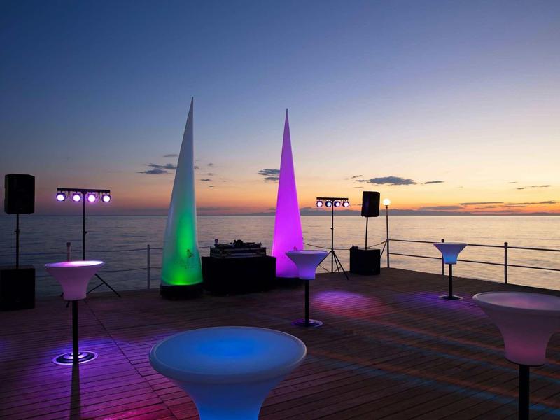 Terrasse am Meer bei Sonnenuntergang mit bunten beleuchteten Lampen und Lautsprechern auf Holzdeck.