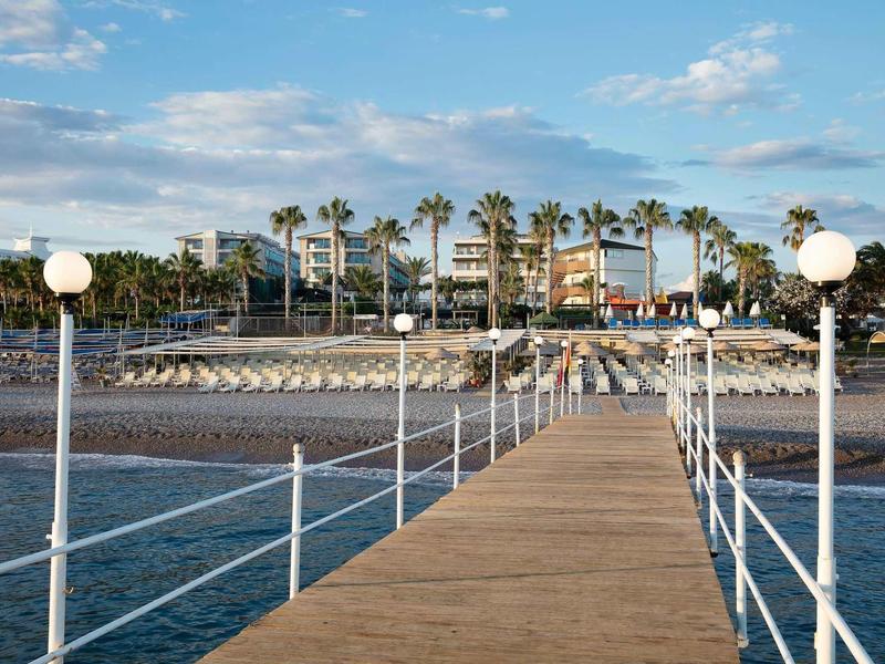 Holzsteg mit weißen Laternen führt zu Palmen und Strandrestaurant unter blauem Himmel.