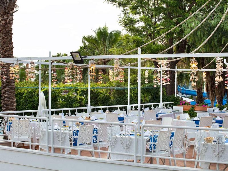 Leerer Außenbereich eines Restaurants mit weißen Tischen, Stühlen und Palmen im Hintergrund.