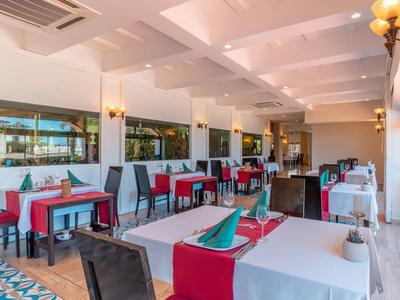 Ristorante luminoso con tavoli bianchi, sedie rosse e grandi finestre con vista sulla piscina.