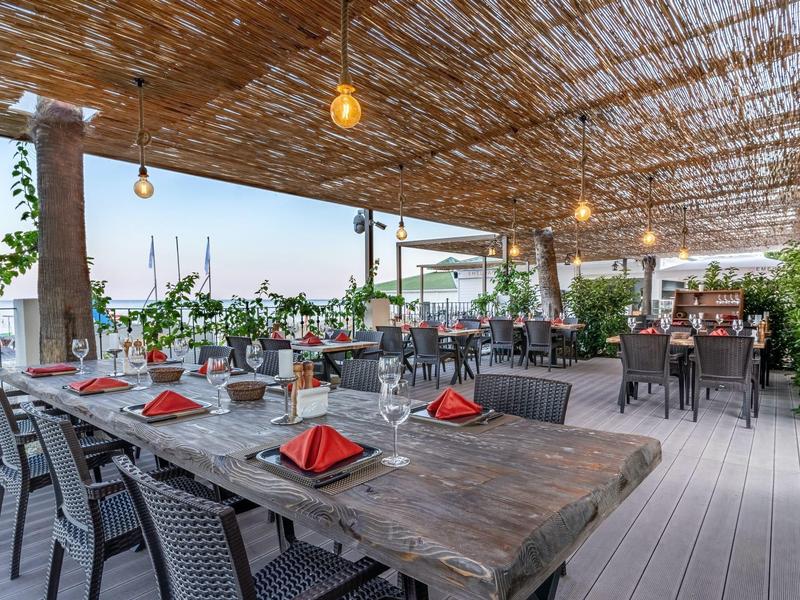 Terrazza esterna coperta con tavoli in legno, tovaglioli rossi e vista sull'acqua.