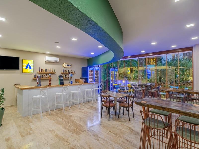 Area bar dell'hotel moderna con mobili chiari, piante verdi e ampia finestra con vista esterna.