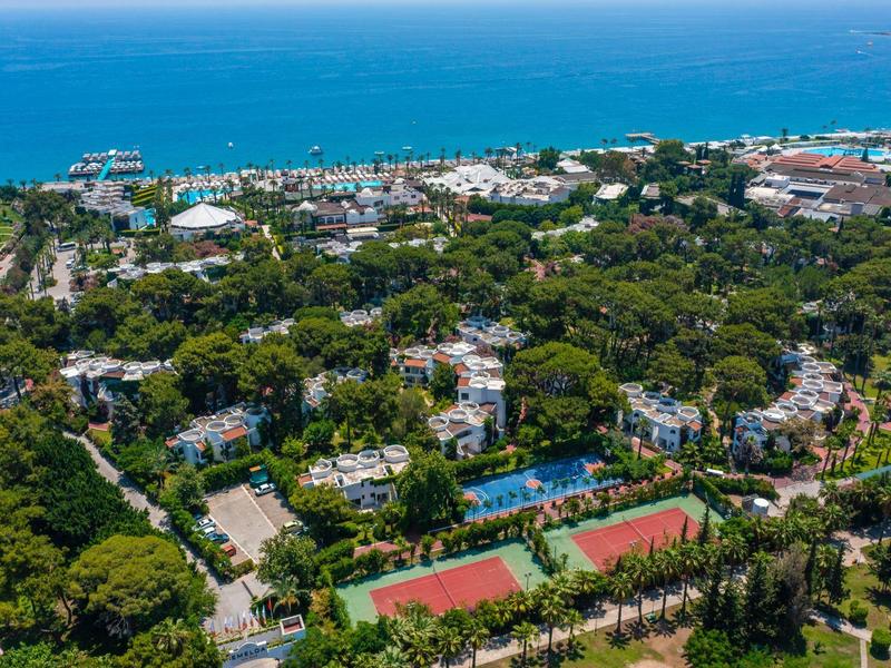 Veduta aerea di un'area costiera boscosa con campi da tennis e mare blu sullo sfondo.