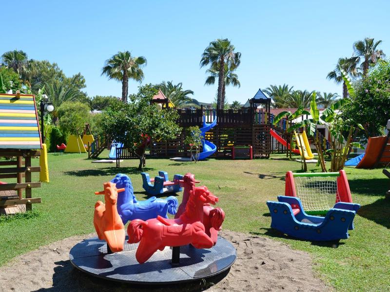 Parco giochi colorato per bambini con altalene, scivoli e strutture da arrampicata su prato verde sotto palme.
