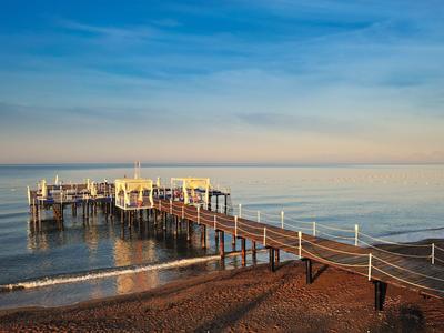 Langer Steg mit Geländern, der über einen Strand ins ruhige Meer führt bei klarem Himmel.