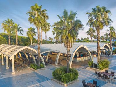 Außenbereich eines Hotels mit geschwungenen Betonbögen, Palmen und Sitzgelegenheiten.