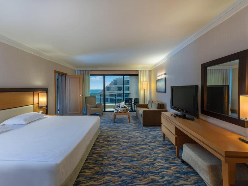 Grande chambre d'hôtel avec lit king-size, télévision, bureau et balcon avec vue sur les bâtiments.