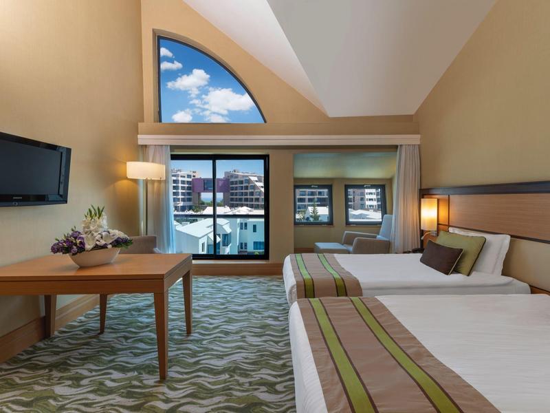 Chambre d'hôtel avec deux lits, bureau et grandes fenêtres avec vue sur la ville.