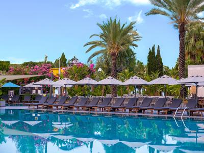 Zwembad met ligstoelen en parasols, omgeven door palmbomen en bloeiende planten.