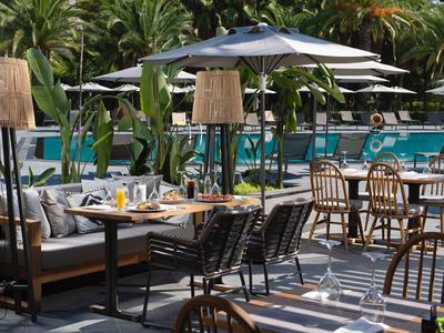 Area ristorante all'aperto con tavoli, sedie e ombrelloni accanto a piante tropicali rigogliose.