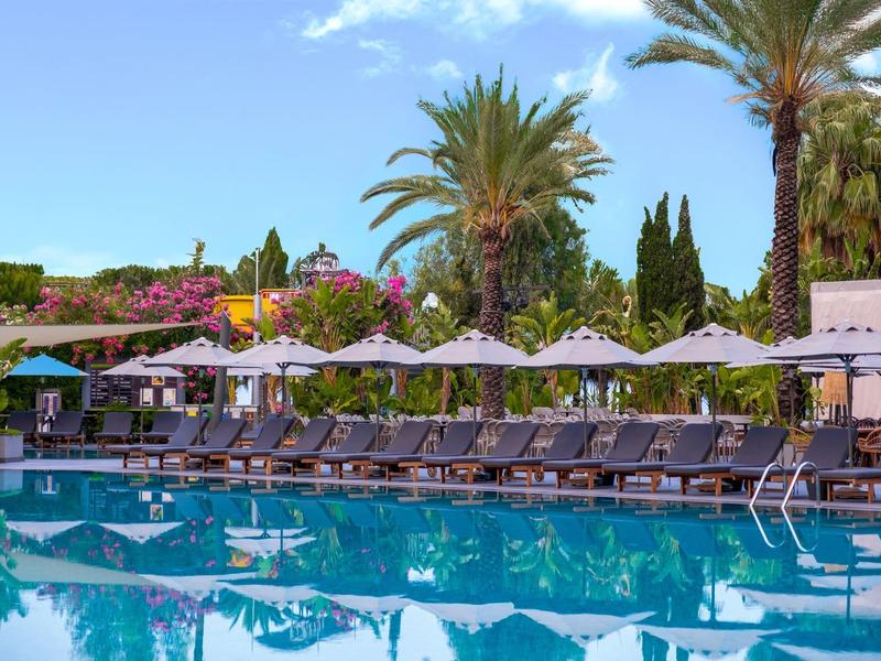 Area piscina con lettini e ombrelloni, circondata da palme e piante.