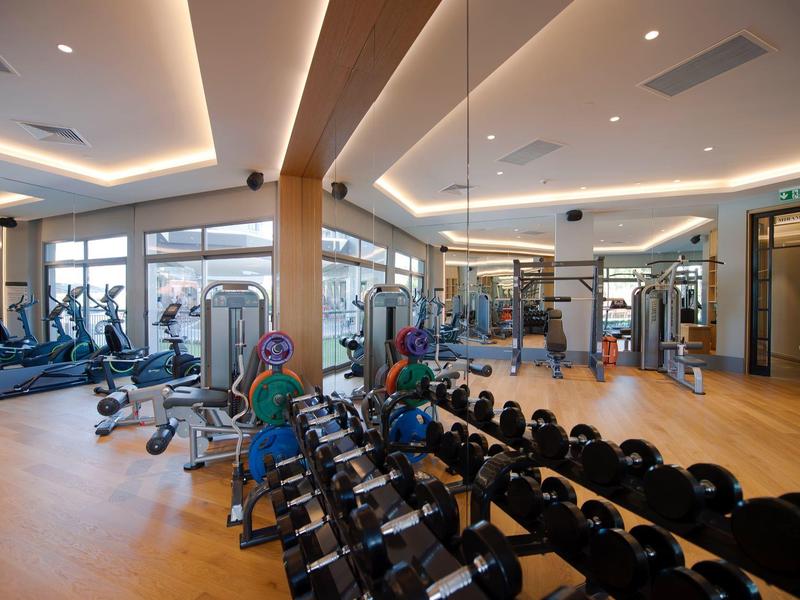 Palestra spaziosa con manubri, attrezzi cardio e grandi finestre con vista esterna.