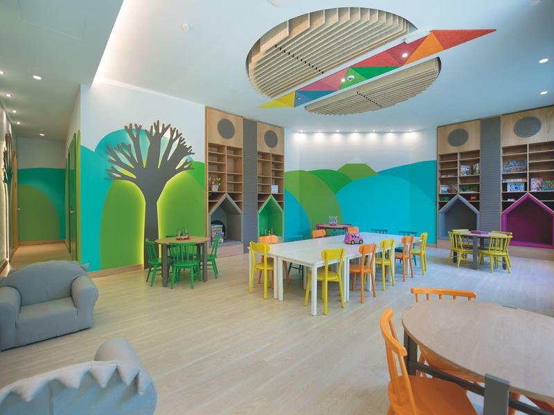 Sala giochi colorata con vari tavoli, sedie e scaffali, pareti decorate con motivi di alberi e montagne.